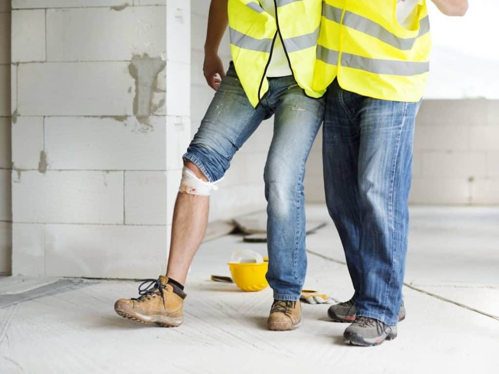 Two construction workers in yellow vests stand indoors; one has a bandaged knee, and a yellow hard hat and tools are on the floor nearby.