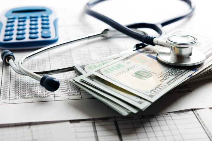 A stethoscope, a stack of hundred-dollar bills, and a blue calculator rest on top of financial documents.