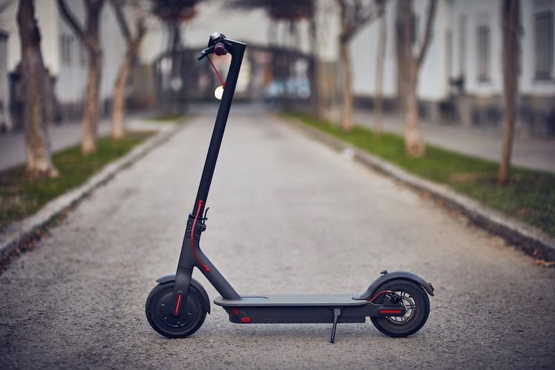 A black and red electric scooter is parked upright on a paved street lined with trees and buildings.