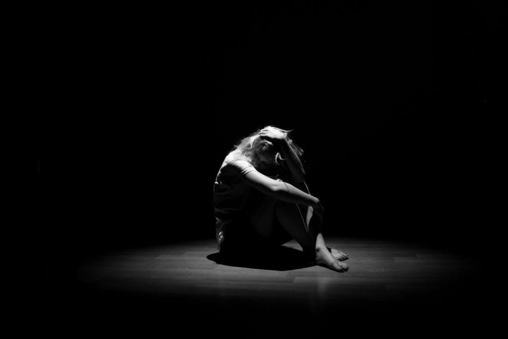 A person sits alone on the floor in a dark room, illuminated by a single spotlight, with their head resting on their knees and one hand in their hair.