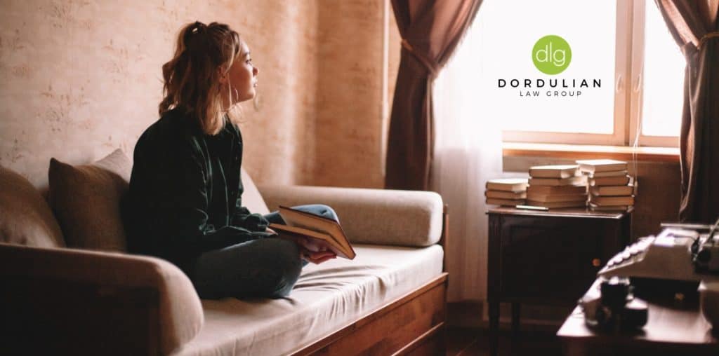 A person sits on a couch holding a book, looking toward a window. A stack of books and a typewriter are on a nearby table. The Dordulian Law Group logo is present.
