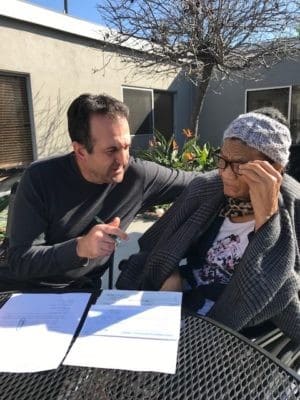 A man sits beside an older woman outdoors, pointing to paperwork on a table as she looks at the documents with a concerned expression.