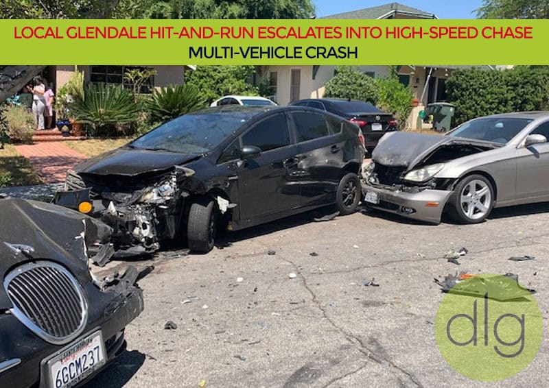 Three heavily damaged cars are stopped on a residential street after a collision; debris is scattered on the road and people are visible in the background.