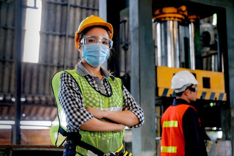 Two workers wearing safety gear, including helmets, masks, and vests, stand in an industrial setting. The worker in the foreground has arms crossed and looks at the camera.