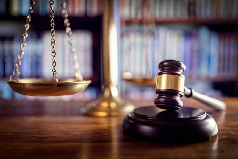 A wooden gavel rests beside brass scales of justice on a desk, with blurred bookshelves in the background.